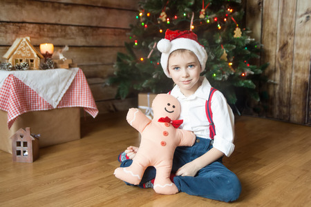Boy in a Christmas hat with gingerbread man toy sitting in front of decorated Christmas tree