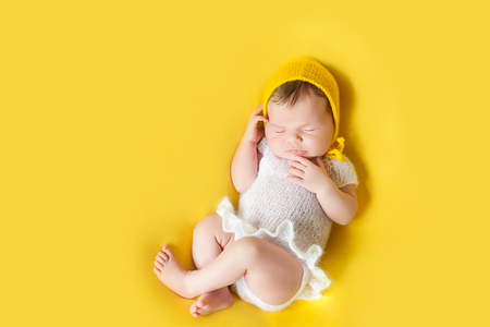 Lovely sleeping newborn baby girl in white knitted fluffy bodysuite and yellow hat lies on her back on a vivid yellow blanketの写真素材