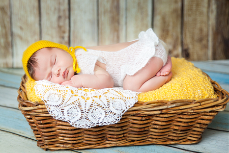 Cute newborn baby girl in a white knit romper and yellow hat sleeping on a yellow blanket and white lace napkin in a basketの写真素材