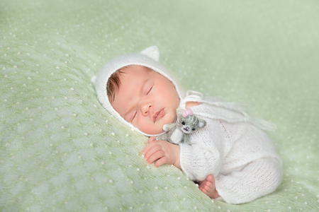 Cute sleeping newborn baby in white knitted fluffy romper with kitten hat with a felt kitten toy lies on the tummy on a light green blanketの写真素材