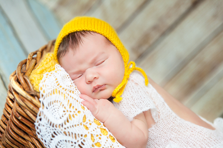 Cute newborn baby girl in a white knit romper and yellow hat sleeping on a yellow blanket and white lace napkin in a basketの写真素材