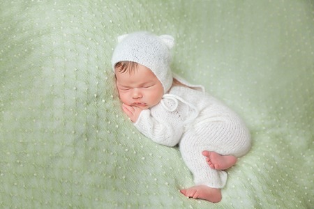 Cute sleeping newborn baby in white knitted fluffy romper with kitten hat lies on the tummy on a light green blanketの写真素材
