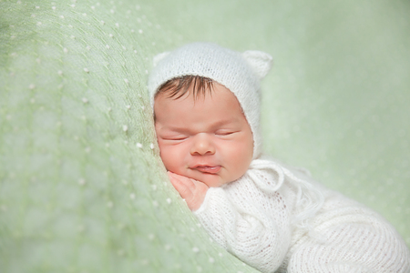 Cute sleeping smiling newborn baby in white knitted fluffy romper with kitten hat lies on the tummy on a light green blanketの写真素材