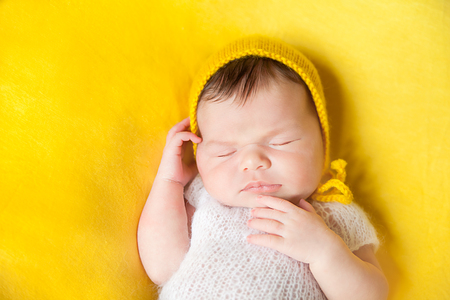 Lovely sleeping newborn baby girl in white knitted fluffy bodysuite and yellow hat lies on her back on a vivid yellow blanketの写真素材