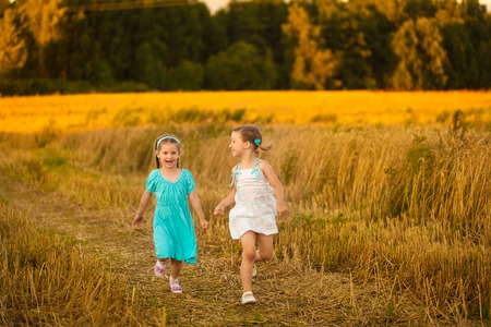 Happy girls in blue and white dress running in wheat field on warm and sunny summer evening. Cute little children in rye field on sunsetの写真素材