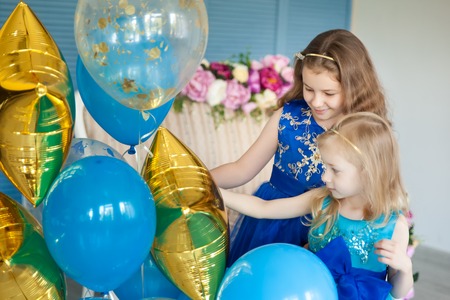 Birthday party in blue and gold collors. Studio session. Fashion girl celebrating her birthday. Two sisters looking at balloons and having fun in the studioの写真素材