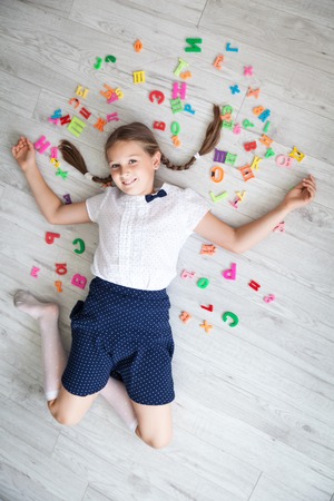 Portrait of a funny school girl in a uniform lying on wooden floor with colorful letters. Back to schoolの写真素材