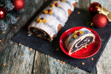 Poppy seed christmas glazed roll under new year tree branch, traditional Chistmas beigli cake on wooden table on black cutting board. Polish makowiec with candied fruits and cranberriesの写真素材