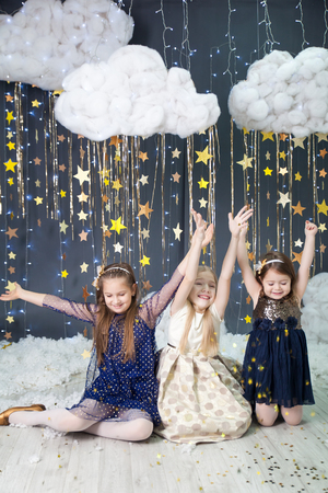 Magic christmas card with three girls in a studio with a gold stars garland, fluffy clouds and lights. Happy girls throw confetti starsの写真素材
