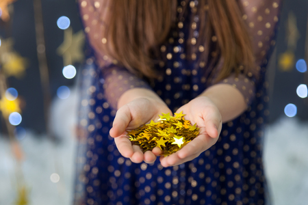 Magic christmas card with a girls hands with confetti stars. Studio with a gold stars garlandand lightsの写真素材
