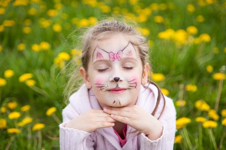 Beautiful young girl with face painting like a kitten dreaming in a dandelion meadow