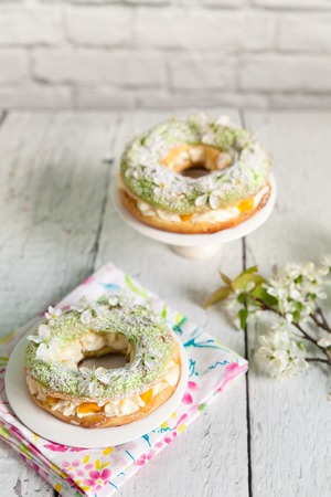 Eclair rings with green craquelin filled with a cream and canned peaches on a white plates on light wooden background with floral napkin. Cherry tree blossom branch. Spring dessert for mother's dayの写真素材