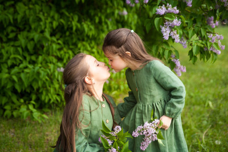 Two sisters in green linen dress have fun in the park with blooming lilacs, enjoy spring and warmth. Beautiful spring garden. Happy childhood. Family, love, peace and happiness conceptの写真素材