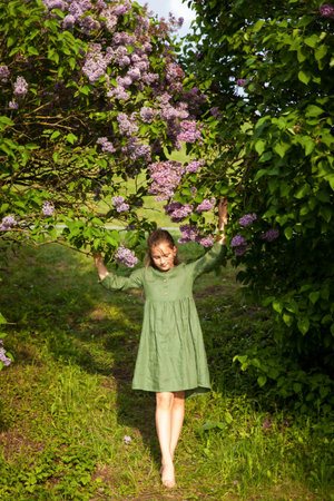 Cute girl in green linen dress has fun in the park with blooming lilacs, enjoys spring and warmth. Beautiful spring garden. Happy childhood, peace and happiness conceptの写真素材