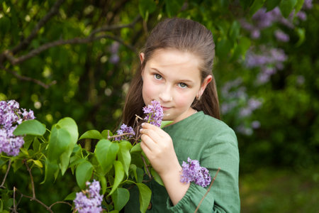 Cute girl in green linen dress has fun in the park with blooming lilacs, enjoys spring and warmth. Beautiful spring garden. Happy childhood, peace and happiness conceptの写真素材
