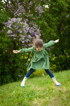 Cute girl in green linen dress has fun in the park with blooming lilacs, enjoys spring and warmth. Beautiful spring garden. Happy childhood, peace and happiness conceptの写真素材