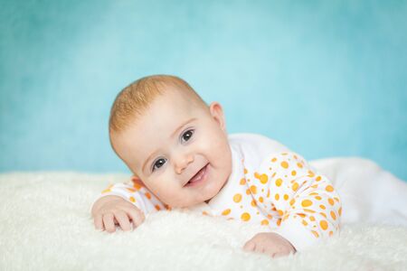 Happy excited 4 months infant baby girl lying on the bed with white fur. Cute girl wearing white...