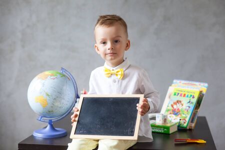 Little preschool boy in a white shirt and with a yellow bow-tie holds a blackboard. back to school concept. Place for text. Distance learning from homeの写真素材