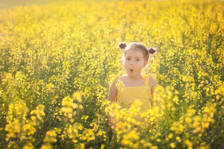 Cute girl in a yellow dress having fun in the field of flowering rape. Nature blooms rape seed field. Summer holidaysの写真素材