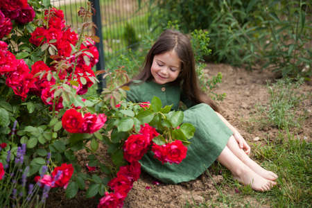 Cute girl in green dress in blossoming rose garden. Cute beautiful lovely child having fun with red roses and lavender in a park on summer dayの写真素材