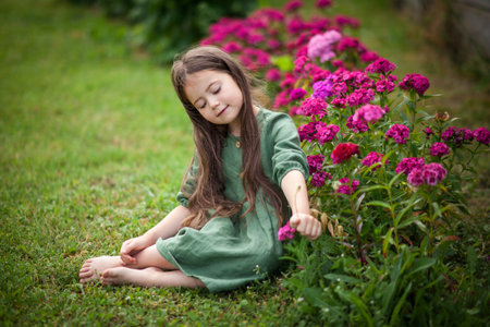 Cute girl in green dress in blossoming garden. Cute beautiful lovely child having fun with pink turkish carnation in a park on summer dayの写真素材