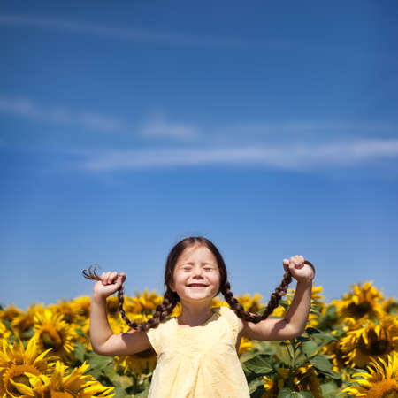 Funny small girl in a yellow dress with pigtails playing in a field of sunflowers. Square image with place for textの写真素材