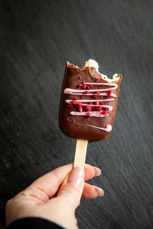 Cheesecake or mousse dessert with freeze-dried raspberries in the form of  ice cream covered with dark chocolate on dark stone tableの写真素材