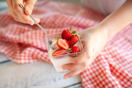 Vanilla panna cotta with strawberry sauce and strawberry berries in a glass cup. Hands holding homemade dessert. Light wooden background with red and white checkered napkinの写真素材