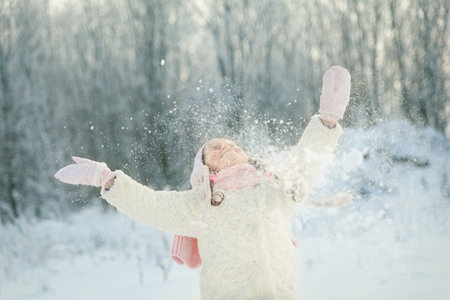 Happy little girl in a white fur coat and a pink knitted scarf plays in a beautiful snowy winter...