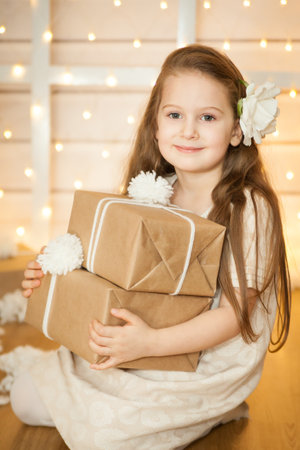 A cute girl holding presents wrapped in craft paper sitting among the winter decorations with Christmas tree and white toys. White Christmas conceptの写真素材