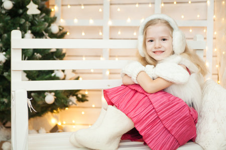 Christmas Eve. Cheerful child in a warm white fur vest, felt boots, mittens and ear flaps sitting on white wooden bench in holiday decorations. White Christmas conceptの写真素材