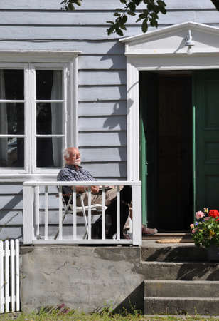 Lærdalsøyri, Norway - August 13, 2011: Old man with white hair and beard relaxing in the sun on the porch of his house. He is sitting on a chair, a newspaper on his lap. A black dog is sitting next to him. The door to his house is open.のeditorial素材