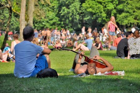 Amsterdam, Netherlands - Juli 23, 2012: Two young men enjoy a summerday in the Vondelpark in Amsterdam. One of them is playing guitar, while the other one is looking at the people on the meadows in the background. Zwei junge Männer genießen einen Sommertのeditorial素材