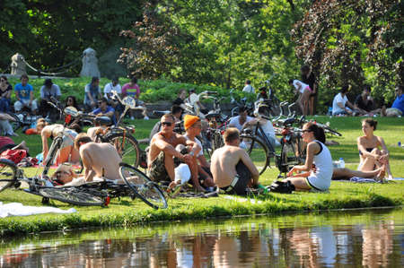 Amsterdam, Netherlands - Juli 26, 2012: Groups of young people enjoying a summerday at Vondelpark. Young men and women sitting and chatting in the gras nearby a pond. Lots a bikes standing and lying arround.のeditorial素材