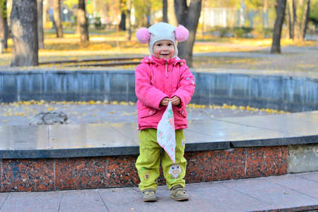 cheerful little girl in a funny hat and a pink jacket with a handkerchief in his hands in a city park. late fall.の写真素材