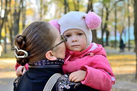little girl in a pink jacket and funny hat feels sad sitting on the hands of his mother. in a city park in the late autumn. mother calms herの写真素材