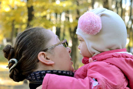 little girl in a pink jacket and funny hat feels sad sitting on the hands of his mother. in a city park in the late autumn. mother calms herの写真素材