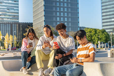 Group of happy teenage friends looking the phone and laughing in a bench in the city street.の写真素材