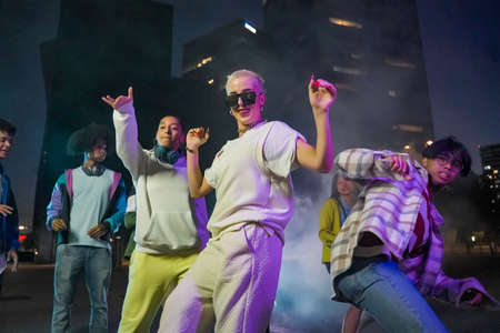 Group of young people dancing in the street at night with modern clothes and colored lights.
