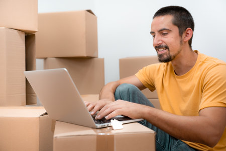 Happy man in his empty new home apartment among the boxes with laptop moving house online trading