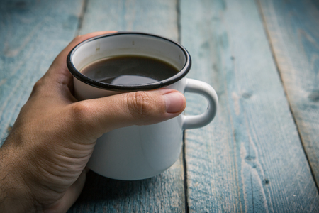 man holding in hand white iron mug with coffee or black tea on an old blue wooden tableの写真素材