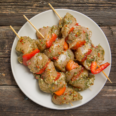skewers of raw chicken meat and vegetables on light plate on dark wooden backgroundの写真素材