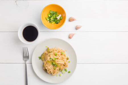 Noodles with bell pepper on plate and garlic cloves and green chopped onion in litle bowl on white wooden backgroundの写真素材