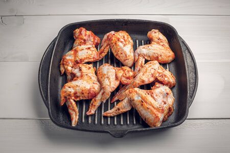 Raw chicken wings marinated in spices and lie in a black cast iron frying pan on a white wooden tableの写真素材