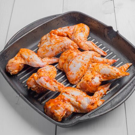 Raw chicken wings marinated in spices and lie in a black cast iron frying pan on a white wooden tableの写真素材