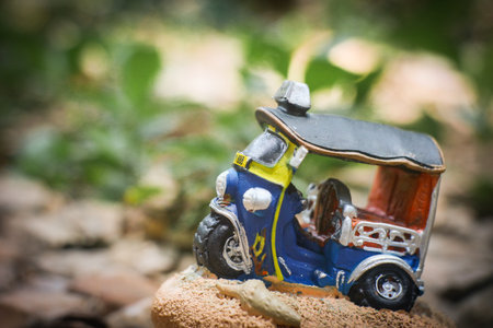 Side view of a colorful clay tuk-tuk model with a flat tire, placed in a rustic countryside scene. Soft-focus trees in the background. Concept of halted journey, nostalgic charm.の写真素材