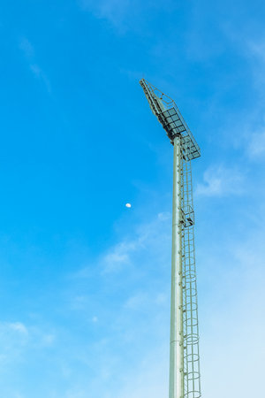 Low angle of stadium spotlight pole with blue sky, clouds, and small crescent moon. Modern sports architecture, outdoor urban vibe.の写真素材
