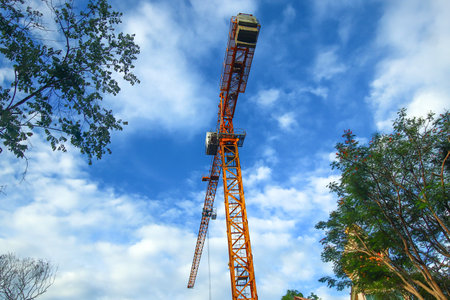 Low angle view of orange crane centered in blue sky with white clouds and green trees. Captures harmony between construction, nature, and sky.の写真素材