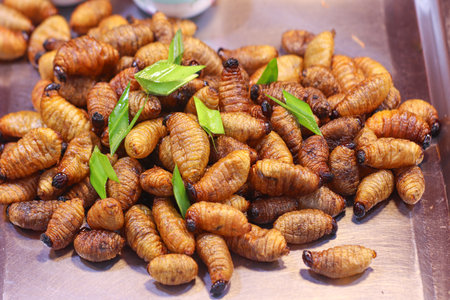 Crispy fried coconut beetle larvae on a tray with pandan garnish, an exotic Thai street food delicacy often enjoyed at night markets and local stalls.の写真素材