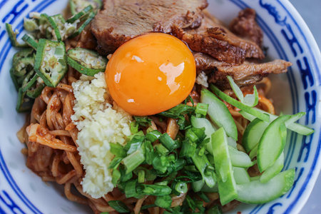 Thai Sichuan-style noodles with roasted red pork, raw egg yolk, and sliced cucumber, representing vibrant and authentic Thai street food.の写真素材